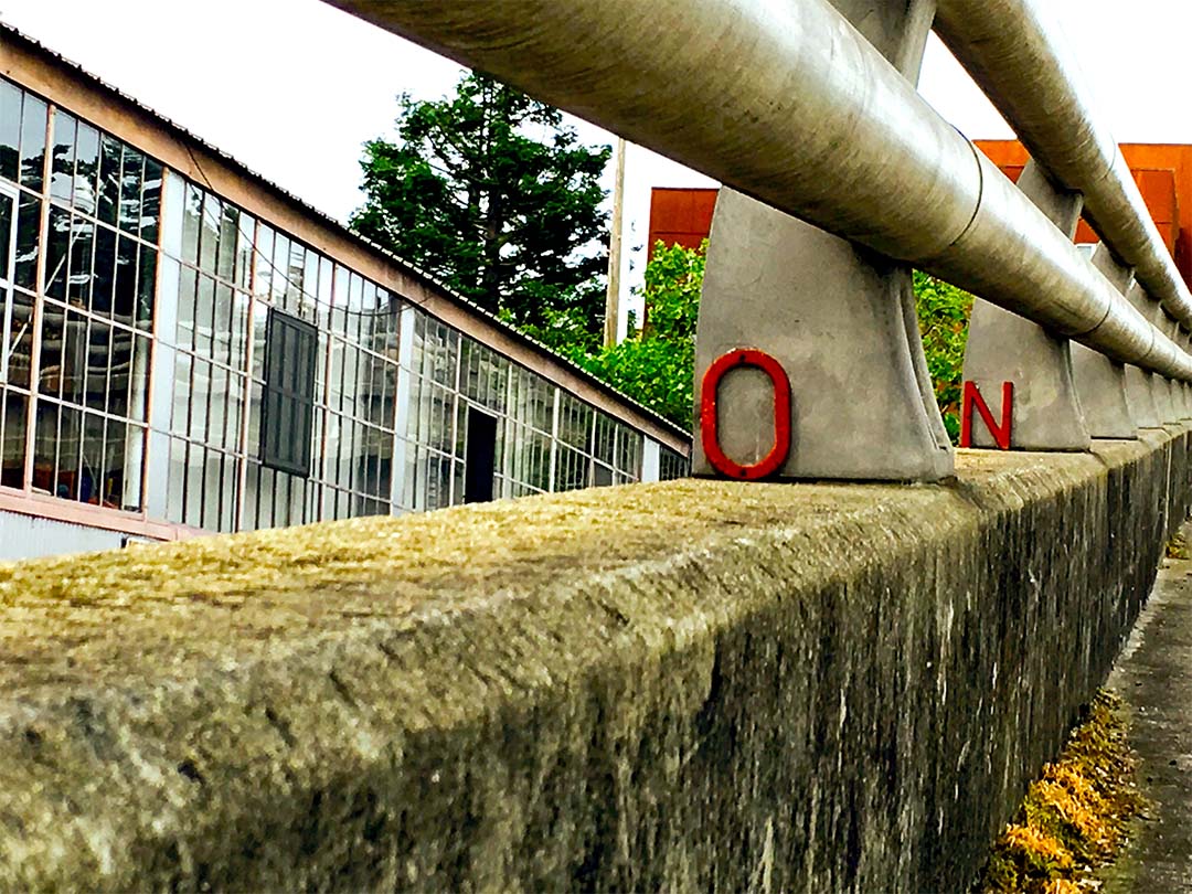 Close shot of a modern bridge&rsquo;s railing, green with moss and natural entrustment. Two bright red letters lean against sequential curved metal columns supporting the railing. In the foreground: O. Against the next support: N.